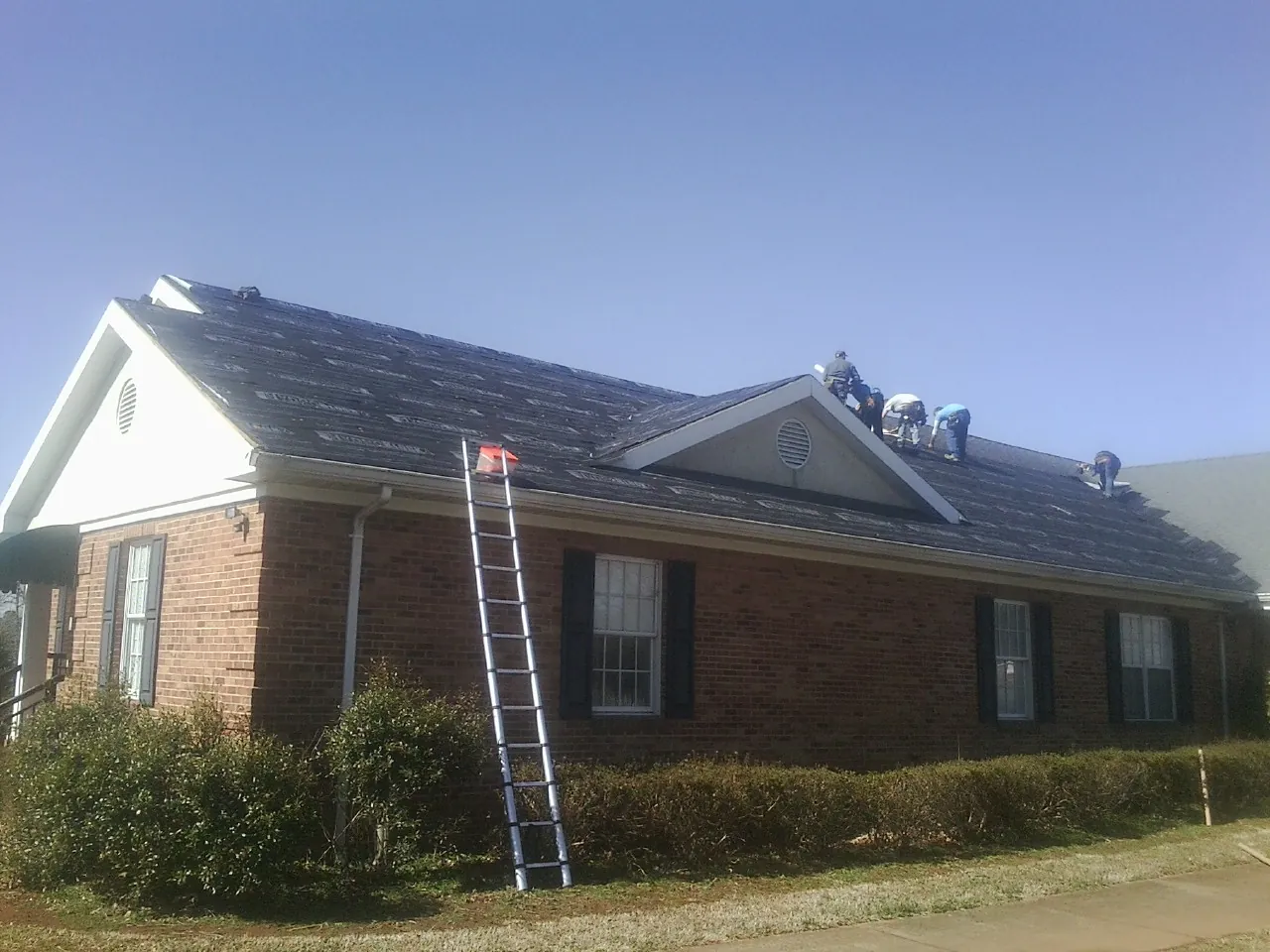 Crew tearing shingles off a brick ranch roof with a ladder set — Metal Roof Repair work by our crew in Lake Mills