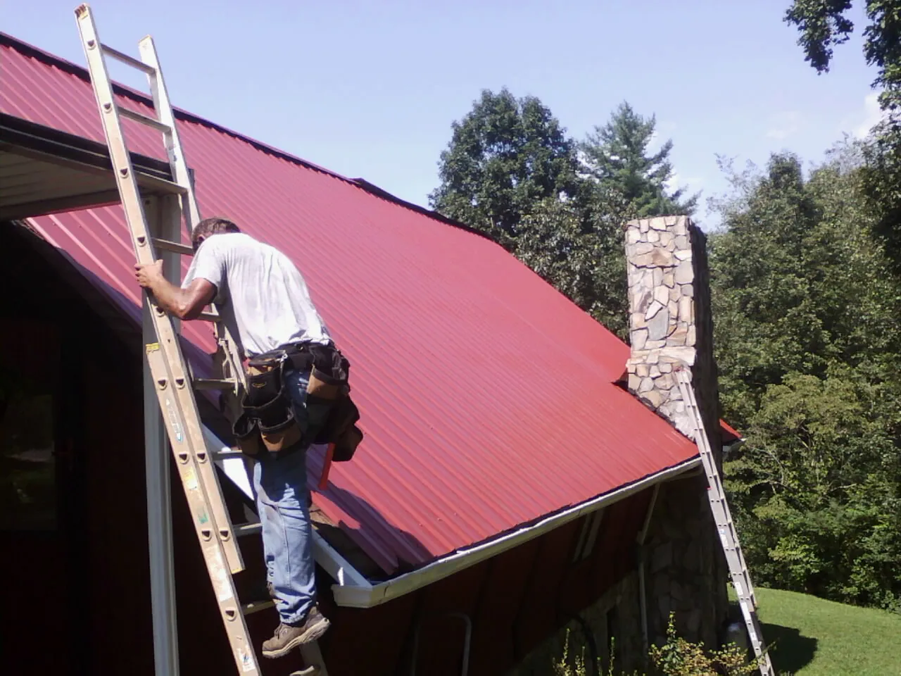 Roofer on a ladder beside a red metal roof and stone chimney — Soffit & Fascia Repair by Roofing Brothers in Lake Mills